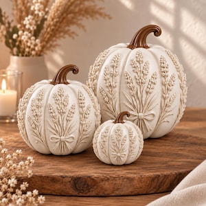 Può includere: Tre zucche decorative color bianco sporco di varie dimensioni, ciascuna caratterizzata da un rilievo scolpito di steli di grano legati con un fiocco. Le zucche hanno corpi lisci e arrotondati e steli curvi di colore marrone scuro.