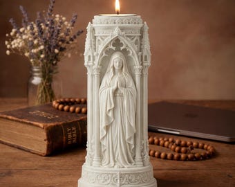 Op de afbeelding: Een witte religieuze kandelaar met een beeld van een vrouw met gevouwen handen in gebed. De kandelaar heeft een sierlijk, gotisch ontwerp met een brandende kaars aan de bovenkant. De kandelaar staat op een houten oppervlak.