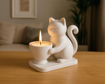 May include: A white ceramic cat-shaped candle holder. The cat is holding a white candle cup with a lit candle. The candle holder sits on a rectangular base. The background includes a wooden table and a blurred living room.