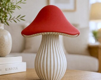 May include: A decorative mushroom-shaped object featuring a smooth, vibrant red cap and a cream-colored, vertically ribbed stem. The piece has a wide, rounded cap that overhangs the textured, cylindrical base, creating a distinct fungal silhouette.