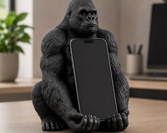 May include: A matte black gorilla figurine serving as a smartphone stand. The gorilla is depicted in a seated, forward-facing position with its hands cupped together to securely hold a black smartphone in a vertical, upright orientation.