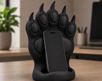 May include: A matte black smartphone stand shaped like a large bear paw with five distinct claws and textured fur detailing. The stand features a central recessed slot designed to hold a rectangular smartphone in an upright, vertical position.