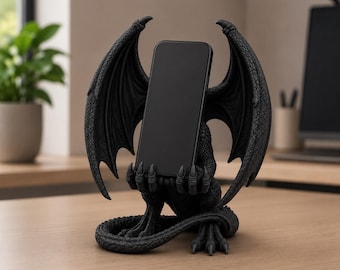 May include: A matte black dragon figurine phone stand featuring a detailed creature with large, textured wings spread wide. The dragon is depicted in a seated position with its clawed hands held forward, securely gripping a black smartphone.