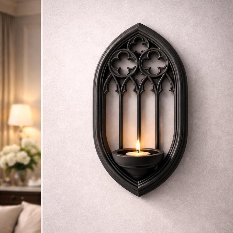May include: Black wall-mounted candle holder with a gothic arch design. The holder features a small, round bowl to hold a lit candle. The frame has a decorative pattern at the top and three vertical arches. The candle flame is visible.