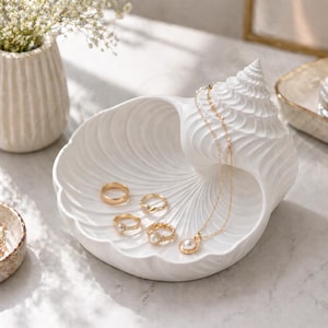 Könnte beinhalten: Eine weiße Schmuckschale aus Keramik in Muschelform mit gewundener Spitze und geripptem Inneren. Die Schale enthält vier goldfarbene Ringe und eine zarte Goldkettenhalskette mit einem kleinen weißen Perlenanhänger in der gewölbten Schale.