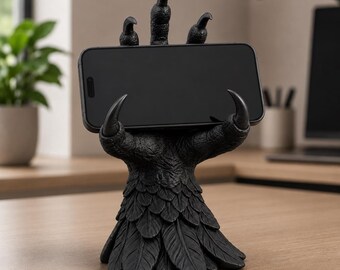 May include: A matte black phone stand shaped like a textured bird claw with sharp talons gripping a horizontal smartphone. The base of the stand features a layered feather design, providing a stable foundation for the device while it is held in place.