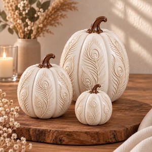 May include: Three off-white decorative pumpkins of varying sizes, each featuring a raised, embossed peacock feather pattern across the surface and a smooth, glossy brown stem. The pumpkins are arranged together as a set of three distinct pieces.