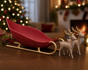 May include: A decorative red sleigh with a leaf-like shape and gold-colored runners, accompanied by two standing beige reindeer figurines featuring gold-colored antlers. The sleigh and reindeer are positioned together as a set.