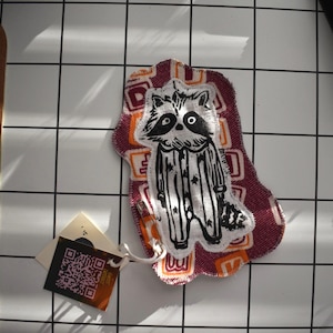 Puede incluir: Parche de tela con una ilustración en blanco y negro de un mapache. El parche está sobre un fondo burdeos con un patrón naranja y blanco. Se adjunta una pequeña etiqueta con un código QR.