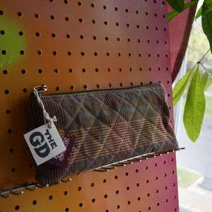 Puede incluir: Un estuche acolchado con un estampado de cuadros en tonos marrón, verde y burdeos, con cierre de cremallera. Una etiqueta con "GD" está adjunta. El estuche descansa sobre una balda metálica contra un panel perforado.