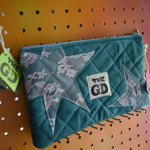 May include: Teal quilted pouch with a star-shaped lace design and a square patch that reads "THE GD". The pouch has a zipper closure and is displayed on a pegboard.