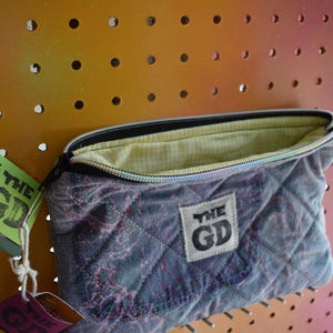 May include: A quilted, zippered pouch in shades of gray and purple, with a yellow and white gingham lining. The pouch has a rectangular label with "THE GD" printed on it. The pouch is hanging on a pegboard.