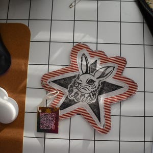May include: A star-shaped fabric coaster with a red and white striped border. The center features a black and white illustration of a rabbit's face within a star. A small tag with a QR code is attached. The coaster is on a white tiled surface.