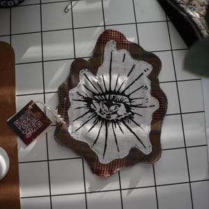 May include: An eye-shaped patch with a black and white eye design on white fabric, surrounded by a brown and plaid border. The patch is irregularly shaped and decorative.