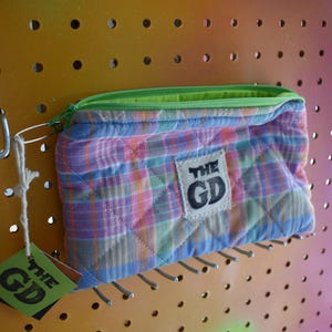 Pode incluir: Uma bolsa acolchoada com padrão xadrez, zíper verde e etiqueta "THE GD". A bolsa é em tons de azul, rosa e verde. Está pendurada em um painel perfurado.
