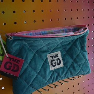 Pode incluir: Embreagem acolchoada verde-azulado com zíper rosa e forro xadrez. A embreagem tem um patch branco com "THE GD" impresso em preto. Duas etiquetas com "GD" estão anexadas. A embreagem é exibida em um painel perfurado.