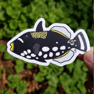 May include: A black and white fish sticker with yellow accents. The fish has a round body with white spots and a yellow stripe along its side. The fish is swimming to the right.