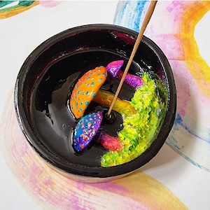 May include: A black ceramic incense holder with colorful mushroom-shaped decorations. The mushrooms have orange, blue, and purple caps with white spots, and red stems. A lit incense stick is placed in the holder, next to a green, textured area.