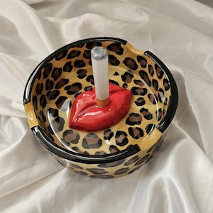 May include: Circular ceramic ashtray with a leopard print pattern. The ashtray has a red lip-shaped cigarette holder. The rim is black, and it holds a lit cigarette.