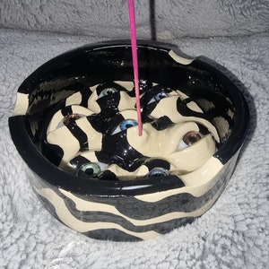Może przedstawiać: Ceramiczny uchwyt na kadzidło z czarno-białym wzorem w paski zebry. Wnętrze przedstawia twarz z wieloma oczami, każde w innym kolorze. Różowa pałeczka kadzidła jest włożona do uchwytu.