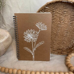 Pode incluir: Um caderno marrom com um design floral branco na capa. O caderno tem uma encadernação em espiral e está fechado.