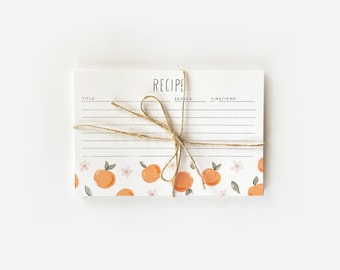 May include: A white recipe card with a brown twine bow. The card has a lined section for writing down a recipe, with headings for "Title", "Serves", and "Time/Temp". The bottom of the card has a repeating pattern of orange blossoms and fruit.