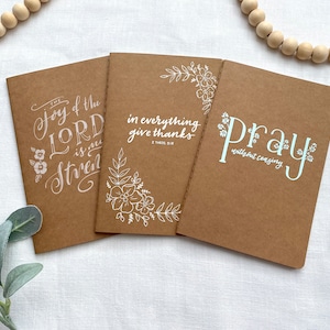 May include: Three brown kraft paper journals with white floral designs and inspirational quotes. The quotes are "The joy of the Lord is my strength", "In everything give thanks", and "Pray without ceasing".