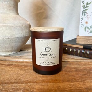 May include: A brown glass candle with a wooden lid. The label reads "Coffee Shop" with coffee, caramel, sugar, and vanilla scents. The candle is 6 oz and handcrafted with natural soy wax. The candle is on a wooden surface.