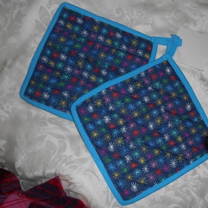 Puede incluir: Dos agarraderas de olla acolchadas azules con un patrón de estrellas de colores y un borde turquesa.