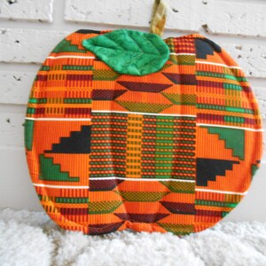May include: A handmade pumpkin-shaped pot holder with a green leaf on top. The pot holder is made of fabric with a colourful African print pattern in orange, green, black, and red.