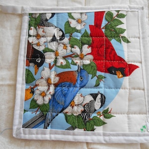 Peut inclure: Un dessous de plat matelassé blanc avec un motif floral bleu, rouge et vert représentant des oiseaux et un cardinal. Le motif est entouré d'une bordure blanche.