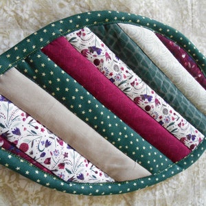Puede incluir: Un posavasos de hoja verde con un diseño acolchado. El posavasos está hecho de diferentes telas en tonos de verde, beige y burdeos. La tela tiene una variedad de patrones, incluyendo estrellas, flores y diseños geométricos.