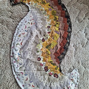 May include: A large, circular, multi-colored patchwork quilt with a spiral design. The quilt is made of various fabrics in shades of brown, beige, yellow, and white. The quilt is edged with a white fabric and has a scalloped edge.