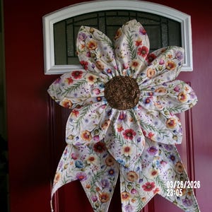 May include: A large, floral-patterned door decoration in the shape of a sunflower. The petals are cream-colored with a pattern of small flowers in red, orange, and purple. The center is brown. The decoration is hanging on a red door.