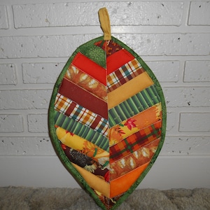 Puede incluir: Un posavasos acolchado con forma de hoja con un borde verde y un diseño de patchwork de telas rojas, naranjas, amarillas y verdes.