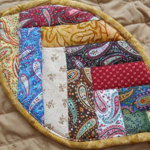 May include: A patchwork leaf-shaped pot holder with a yellow border. The pot holder is made of various fabrics with paisley patterns in shades of red, blue, brown, and white.