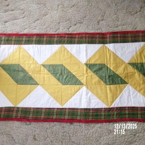 Op de afbeelding: Een rechthoekige, gewatteerde tafelloper met een herhalend patroon van gele en groene ruiten op een witte achtergrond. De rand is een rood en groen geruit patroon. De loper is waarschijnlijk handgemaakt.