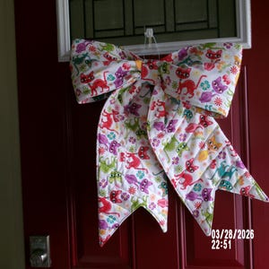 Peut inclure: Un grand nœud en tissu matelassé avec un fond blanc et un motif de chats colorés. Le nœud est accroché à une porte rouge. Le motif comprend des chats rouges, verts, violets et jaunes, avec des fleurs. La date et l'heure sont visibles sur la porte.