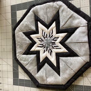 May include: A handmade, octagonal trivet featuring a starburst design. The trivet is primarily gray, black, and white, with a central star pattern and a black border. The trivet measures approximately 9 inches in diameter.