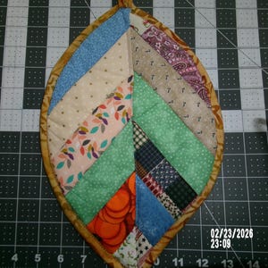 May include: A leaf-shaped quilted potholder with a variety of fabric patterns and colors, including blue, beige, green, and orange. The edges are trimmed with a gold-colored fabric. The date and time 02/23/2026 23:09 are visible in the bottom right corner.