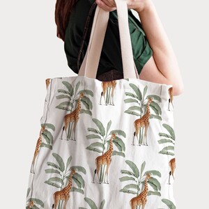 May include: White tote bag featuring a repeating pattern of giraffes and green banana leaves. The bag has beige straps and is held by a person. The giraffe design is in shades of brown and tan.