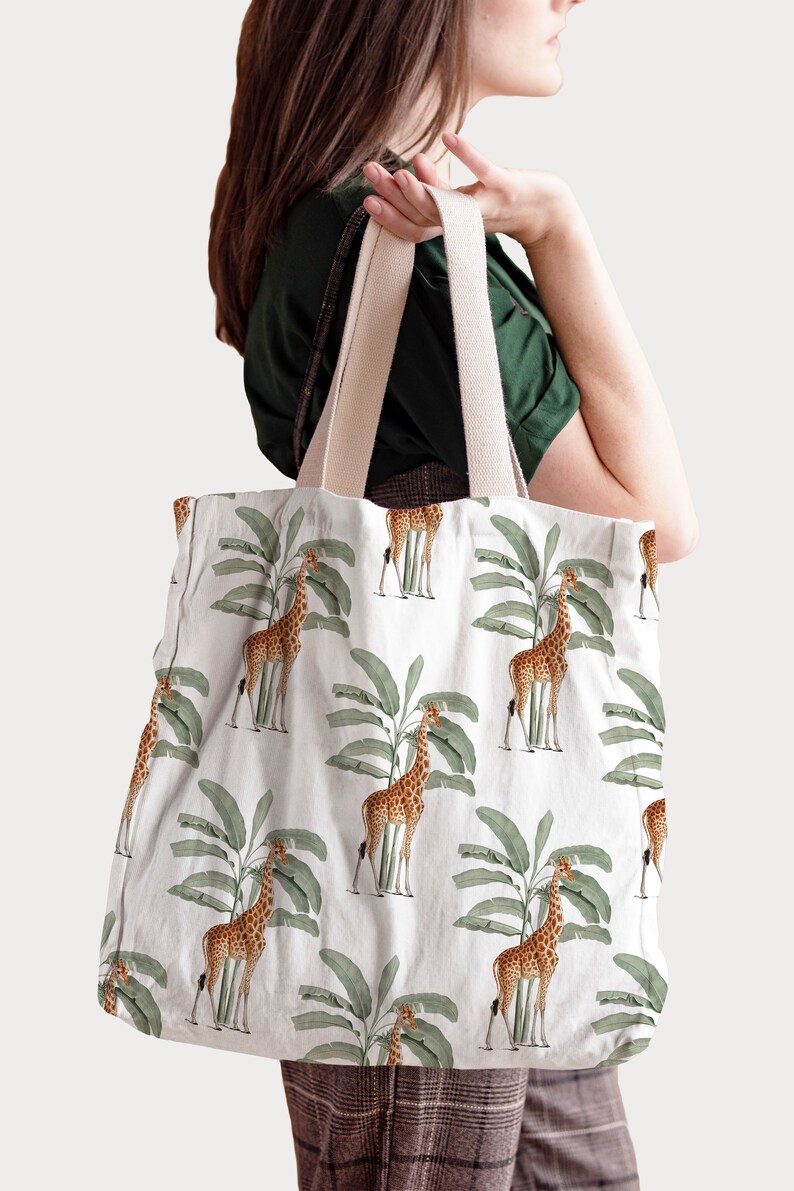 May include: White tote bag featuring a repeating pattern of giraffes and green banana leaves. The bag has beige straps and is held by a person. The giraffe design is in shades of brown and tan.