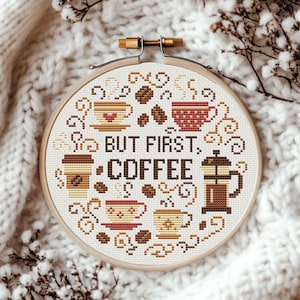 May include: A round embroidery hoop with a cross-stitch design featuring coffee cups, coffee beans, and the text "But First, Coffee".