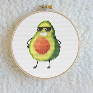 May include: A cross-stitch embroidery hoop with a cartoon avocado wearing sunglasses. The avocado is green with a brown pit and has a smiling face.