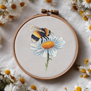 Puede incluir: Un bordado de punto de cruz de un abejorro sobre una margarita, en un aro de madera. La abeja tiene rayas negras y amarillas. La margarita tiene pétalos blancos y un centro amarillo. El fondo es blanco, con margaritas esparcidas.