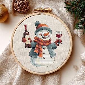 Op de afbeelding: Kruissteekborduurwerk in een houten hoepel met een sneeuwpop die een wijnfles en een glas rode wijn vasthoudt. De sneeuwpop draagt een hoed, sjaal en jas. Het ontwerp staat tegen een neutrale stoffen achtergrond, met kerstversieringen.