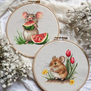 Op de afbeelding: Twee borduurringen met kruissteek. Eén toont een muis die watermeloen eet, de andere een muis met bloemen. De ringen zijn rond, met een lichtgekleurde stoffen achtergrond. De muis is bruin en wit, met roze oren. De watermeloen is rood en groen.