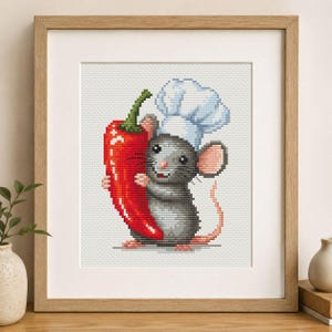Könnte beinhalten: Ein gerahmtes Kreuzstich-Stickbild, das eine graue Maus mit weißer Kochmütze zeigt, die eine große rote Chilischote hält. Das Motiv ist im Pixel-Stil auf weißem Stoff gearbeitet und befindet sich in einem hellen Holzrahmen.