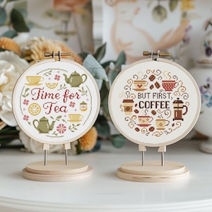 May include: Two cross-stitch hoops on wooden stands. One says "Time for Tea" with teacups, teapots, and flowers. The other says "But First, Coffee" with coffee cups and a French press. Both are in wooden hoops.