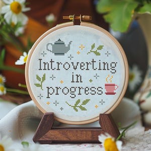 May include: A cross-stitch embroidery hoop with the text "Introverting in progress." The design features a teapot, a teacup, and floral accents. The embroidery is displayed on a wooden stand, surrounded by daisies.